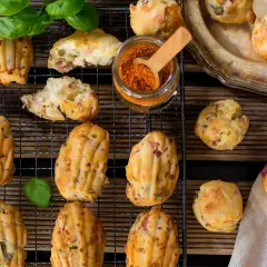Des madeleines salées originales, garnies de lardons et d'herbes, sur une grille avec un pot d'épices et des feuilles de basilic.