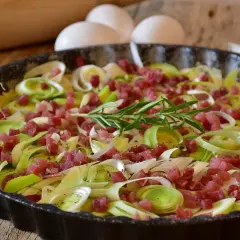 Tarte aux poireaux et lardons, une idée savoureuse pour accompagner des poireaux. Des œufs et un rouleau à pâtisserie sont visibles en arrière-plan.