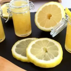 Trois pots de confiture de citron à la mentonnaise, accompagnés de citrons frais coupés et d'une spatule en bois.
