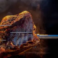 Un morceau de viande assaisonné tourne lentement sur une broche, dégageant une fumée alléchante. La cuisson à la broche promet un délice savoureux.