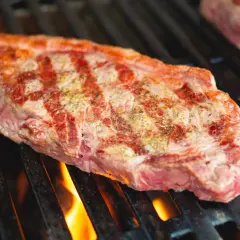 Un steak de basse côte, assaisonné, grésille sur un barbecue. Les flammes dansent sous la grille, promettant une cuisson parfaite.