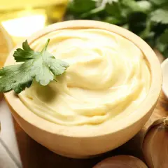Mayonnaise onctueuse dans un bol en bois, garnie de persil. Citrons, ail et moutarde à proximité. La clé pour ne pas rater une mayonnaise maison.