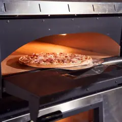 Une pizza garnie est enfournée dans un four à pizza professionnel. La pelle à pizza glisse sous la pâte dorée, prête à être cuite.