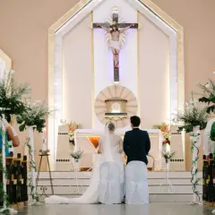 Le déroulement du mariage à l'église commence avec le couple devant l'autel, sous un crucifix. Les invités sont assis dans les bancs décorés de fleurs.