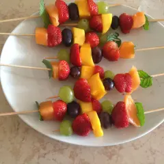 Brochettes de fruits originales : fraises, melon, mangue, raisins et menthe sur des bâtonnets en bois, présentées sur une assiette blanche.