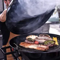 Un homme soulève le couvercle d'un barbecue fumant, révélant une sélection alléchante de quelle viande au tourne broche : saucisses, steaks, lard et légumes grillés.