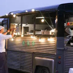Un food truck illuminé de guirlandes lumineuses, parfait pour une idée food truck mariage. Un serveur sert un plat.