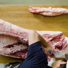 Un boucher démontre comment découper un cochon entier sur une planche en bois. Des morceaux de gras sont déjà séparés.