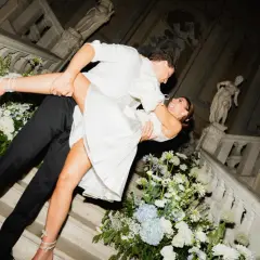 Un couple s'embrasse passionnément lors de leur mariage chez soi, entouré de fleurs blanches et bleues sur un escalier orné de statues.