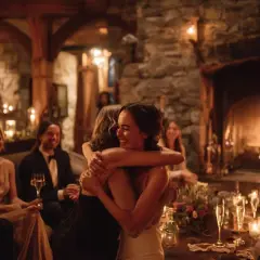 Deux femmes s'enlacent joyeusement, entourées d'invités lors d'une réception chaleureuse. L'ambiance intime et les bougies suggèrent qu'elles sont en train d'organiser un petit mariage.