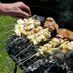 Une main vérifie des brochettes de poulet et des steaks sur un barbecue rustique.