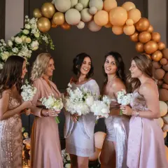 Cinq femmes souriantes, entourées de fleurs et de ballons, se préparent pour un mariage. Elles tiennent des bouquets, prêtes pour la cérémonie.