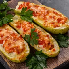 Trois courgettes farcies, façon pizza, gratinées au fromage et garnies de tomates, sur une planche en bois, décorées de persil frais.
