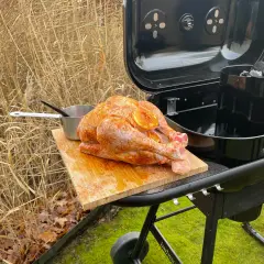 Un dindon assaisonné prêt pour un barbecue de Noël, posé sur une planche à découper à côté d'un barbecue ouvert.