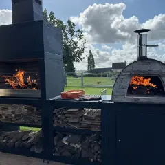 Un barbecue et un four à pizza en pierre, tous deux allumés, prêts pour un repas. Idéal pour savoir comment faire un barbecue sans charbon, avec du bois stocké en dessous.