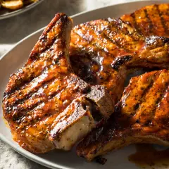 Trois côtes de porcelet grillées, nappées d'une sauce caramélisée et parsemées de poivre noir, sur une assiette blanche.