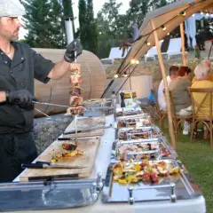 Un chef prépare un menu barbecue mariage. Des brochettes de viande et des légumes sont présentés dans des plats chauffants, tandis que des invités dînent sous une tente décorée de guirlandes lumineuses.