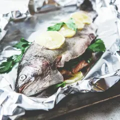 Une truite entière, prête à être cuite en papillote, garnie de rondelles de citron et de persil frais, sur une feuille d'aluminium froissée.