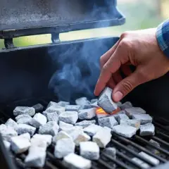 Une main allume des briquettes de charbon dans un barbecue, de la fumée s'échappe. La question est : combien de temps pour allumer un barbecue ?