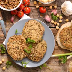 Trois galettes de lentilles, parfaites pour une recette de steak végétarien, sont présentées avec des pains à burger, des tomates et de l'ail sur fond de bois.