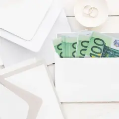 Enveloppes blanches, fleurs et billets de 100€ pour savoir comment offrir de l'argent pour un mariage. Bague de fiançailles sur une soucoupe.