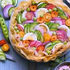 Un plat d'été raffiné : une tarte salée garnie de concombre, radis, œufs durs, tomates cerises, petits pois et oignons rouges, le tout sur un lit de crème rose.