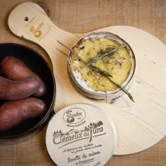 Un crémeux du Jura au four, doré et fondant, avec des pommes de terre rouges dans un bol. Une planche en bois ornée du logo "Crémeux du Jura" complète la scène.