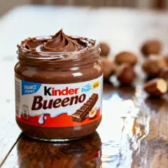 Un pot de pâte à tartiner Kinder Bueno, crémeuse et onctueuse, sur une table en bois. Des noisettes sont disposées en arrière-plan.