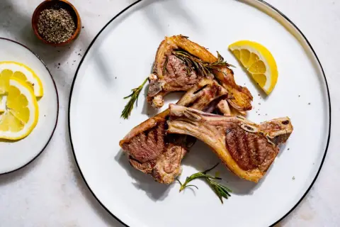 Trois côtelettes d'agneau grillées, garnies de romarin frais, avec des quartiers de citron et du poivre concassé en accompagnement.