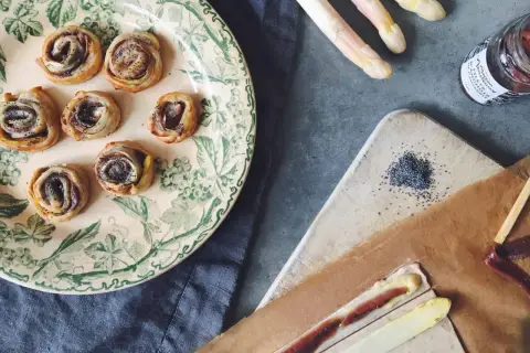 Petits roulés feuilletés à l'asperge blanche, parfaits pour un apéritif. Des asperges blanches, des graines de pavot et une touche de anchois complètent la scène.