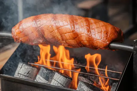 Un jambon à la broche dore au-dessus de flammes vives. La viande, marquée de croisillons, est rôtie lentement pour une saveur incomparable.