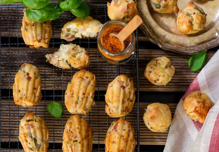 Des madeleines salées originales, garnies de lardons et d'herbes, sur une grille avec un pot d'épices et des feuilles de basilic.