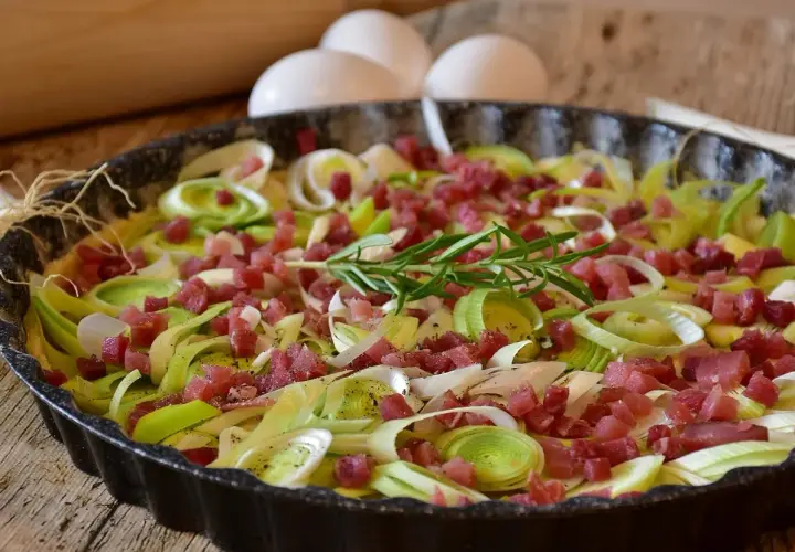 Tarte aux poireaux et lardons, une idée savoureuse pour accompagner des poireaux. Des œufs et un rouleau à pâtisserie sont visibles en arrière-plan.