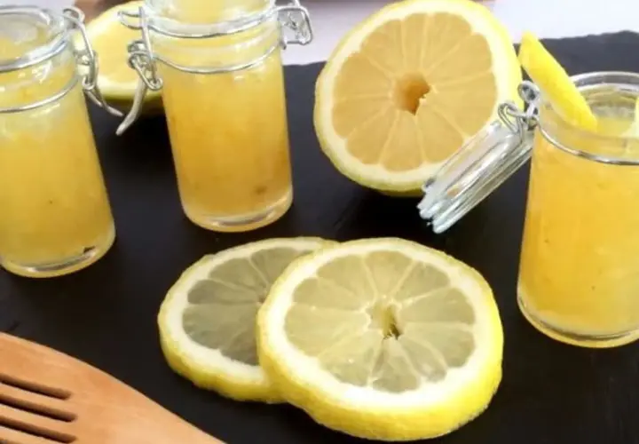 Trois pots de confiture de citron à la mentonnaise, accompagnés de citrons frais coupés et d'une spatule en bois.