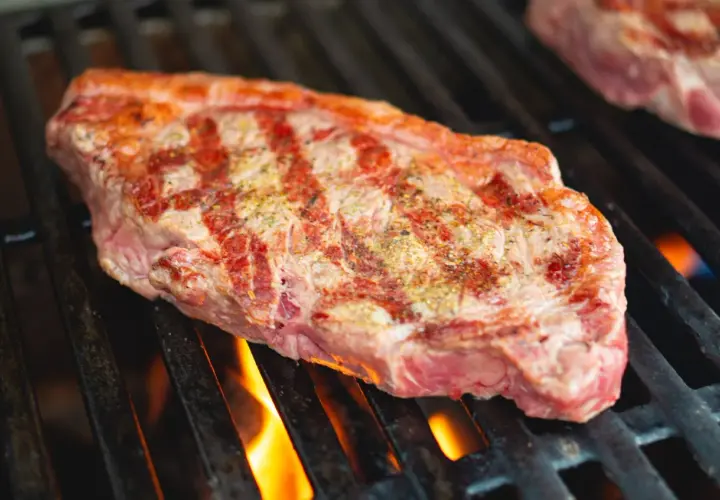 Un steak de basse côte, assaisonné, grésille sur un barbecue. Les flammes dansent sous la grille, promettant une cuisson parfaite.