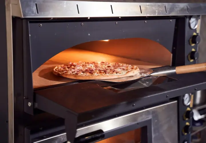 Une pizza garnie est enfournée dans un four à pizza professionnel. La pelle à pizza glisse sous la pâte dorée, prête à être cuite.