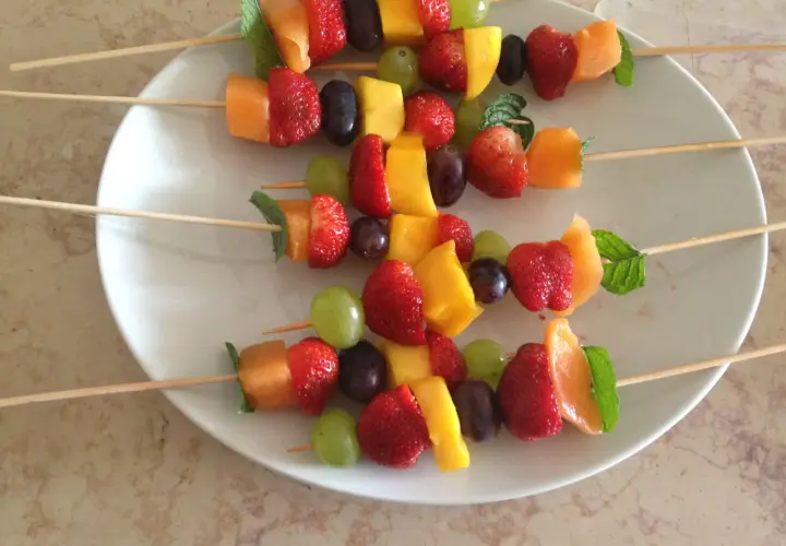 Brochettes de fruits originales : fraises, melon, mangue, raisins et menthe sur des bâtonnets en bois, présentées sur une assiette blanche.