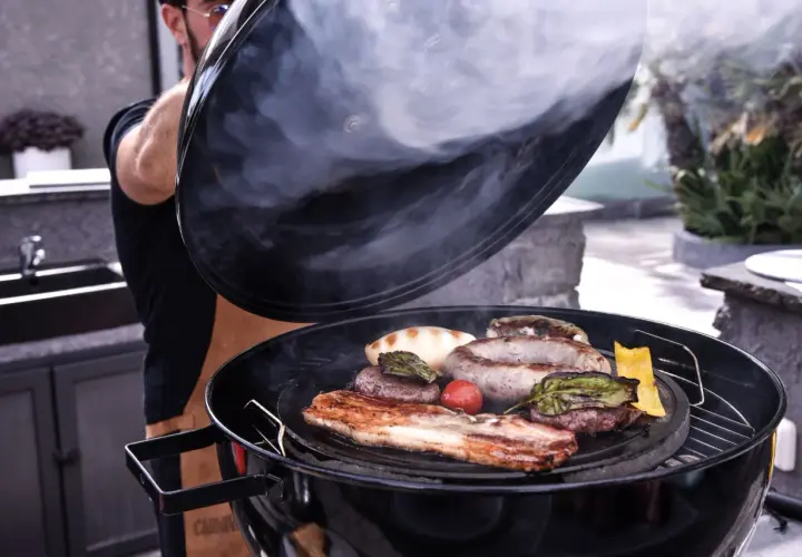 Un homme soulève le couvercle d'un barbecue fumant, révélant une sélection alléchante de quelle viande au tourne broche : saucisses, steaks, lard et légumes grillés.