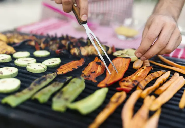 Des légumes accompagnement barbecue grillent sur le feu : courgettes, poivrons et carottes. Un homme les retourne avec une fourchette.