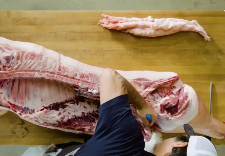Un boucher démontre comment découper un cochon entier sur une planche en bois. Des morceaux de gras sont déjà séparés.
