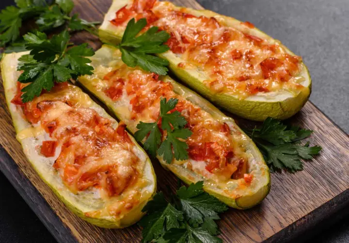 Trois courgettes farcies, façon pizza, gratinées au fromage et garnies de tomates, sur une planche en bois, décorées de persil frais.