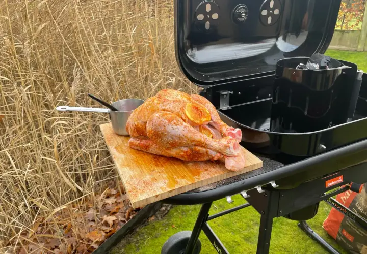 Un dindon assaisonné prêt pour un barbecue de Noël, posé sur une planche à découper à côté d'un barbecue ouvert.