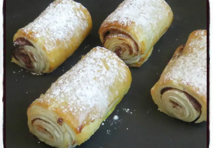 Quatre croissants au chocolat en pâte feuilletée, saupoudrés de sucre glace, prêts à être dégustés.