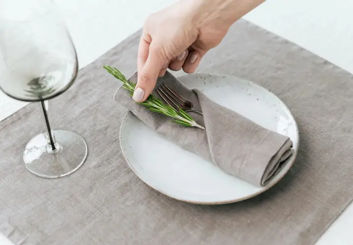 Une main dépose une branche de romarin sur un pliage de serviette mariage, sur une assiette blanche.