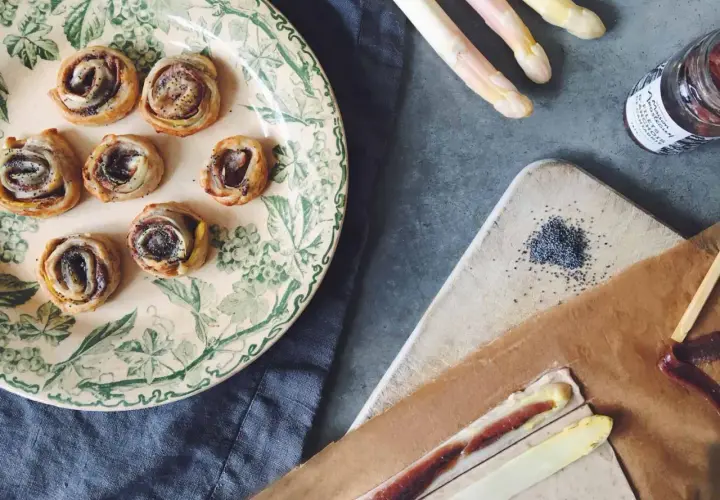Petits roulés feuilletés à l'asperge blanche, parfaits pour un apéritif. Des asperges blanches, des graines de pavot et une touche de anchois complètent la scène.