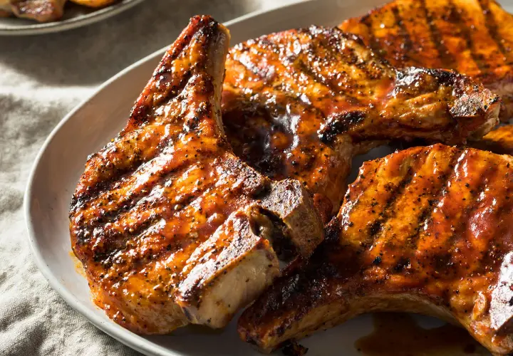 Trois côtes de porcelet grillées, nappées d'une sauce caramélisée et parsemées de poivre noir, sur une assiette blanche.