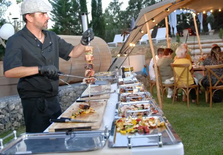 Un chef prépare un menu barbecue mariage. Des brochettes de viande et des légumes sont présentés dans des plats chauffants, tandis que des invités dînent sous une tente décorée de guirlandes lumineuses.