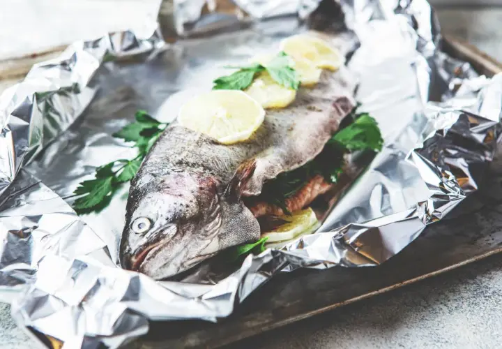 Une truite entière, prête à être cuite en papillote, garnie de rondelles de citron et de persil frais, sur une feuille d'aluminium froissée.