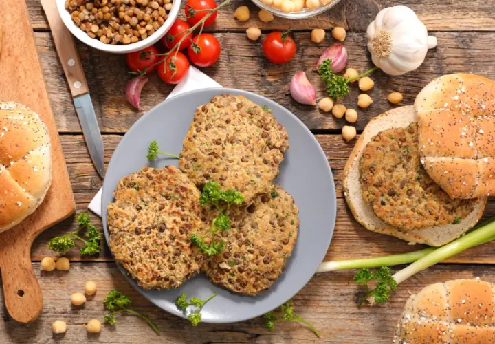 Trois galettes de lentilles, parfaites pour une recette de steak végétarien, sont présentées avec des pains à burger, des tomates et de l'ail sur fond de bois.