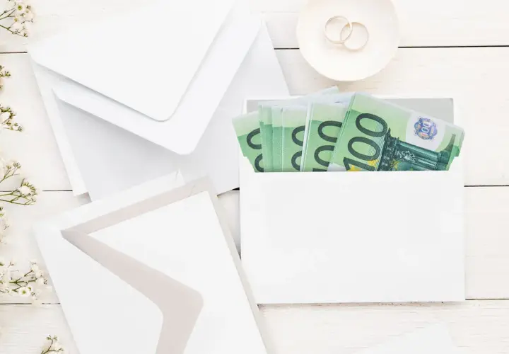 Enveloppes blanches, fleurs et billets de 100€ pour savoir comment offrir de l'argent pour un mariage. Bague de fiançailles sur une soucoupe.